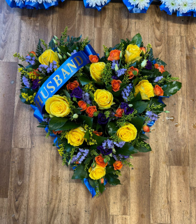 Forever - A stunning heart-shaped funeral tribute featuring a beautiful combination of sunshine yellow, warm orange and rich purple tones, complemented by lush greenery. Finished with elegant blue ribbon edging and a personalised HUSBAND ribbon, this touching arrangement is a loving tribute to a cherished husband, symbolising everlasting love and a lifetime of precious memories together.